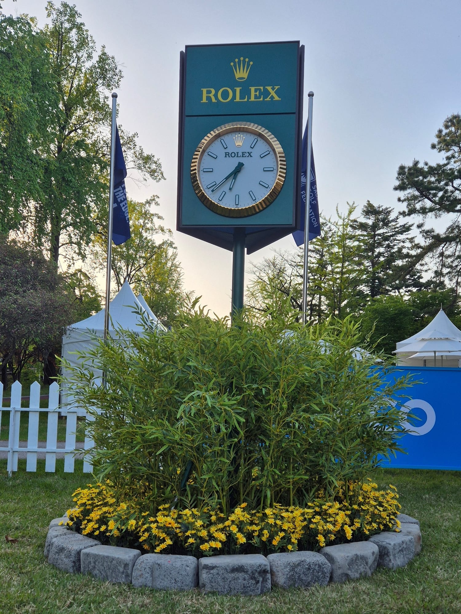 고양시 뉴코리아CC — 골프장 ROLEX 시계탑 아래 인조 대나무와 노란 꽃으로 채운 원형 화단, 석재로 경계를 두른 모습